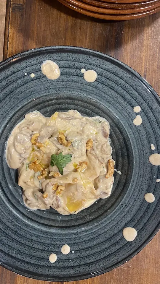Pasta rellena en salsa cremosa con nueces por encima, vista desde arriba en un plato de gres oscuro, con gotas de salsa alrededor y una pila de cazuelas de barro al fondo de la mesa.