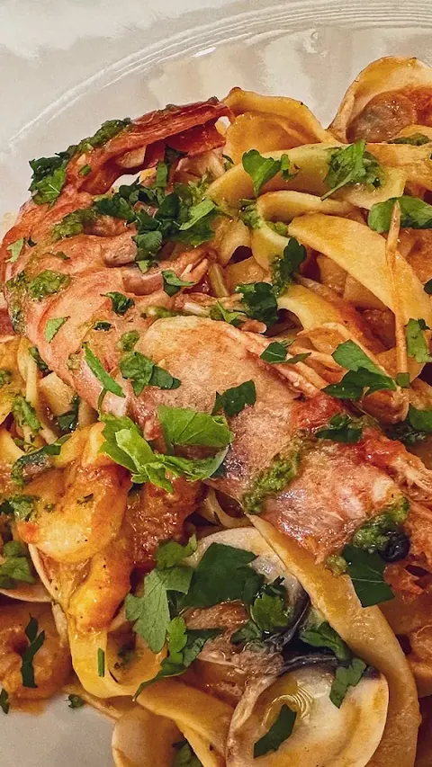 Tagliatelle con gambas enteras con cabeza, almejas abiertas en su concha y mucho perejil picado por encima, de muy cerca, en un plato blanco.
