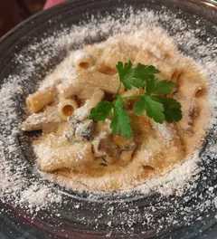 Pasta corta y rayada en salsa cremosa, con una rama de perejil encima y una lluvia de queso rallado que llega hasta el borde, en un plato de gres oscuro.