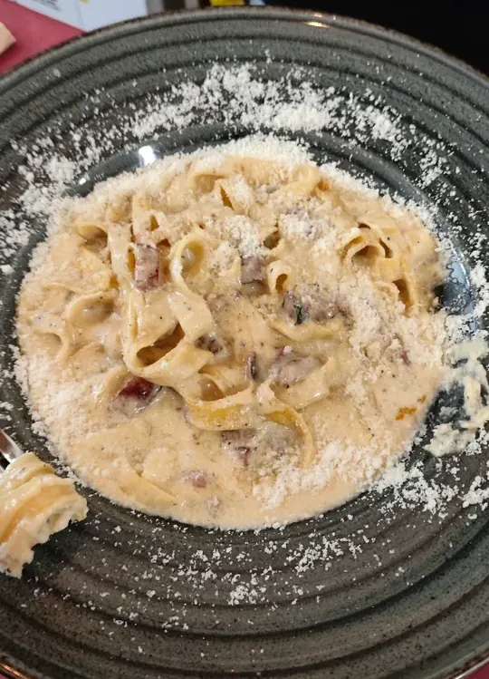 Tagliatelle en una salsa cremosa muy abundante, con dados de bacón, pimienta negra y mucho queso rallado, en un plato de gres oscuro con anillos torneados.