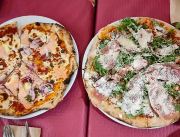 Dos de nuestras pizzas vistas desde arriba, en platos blancos sobre el mantel granate: una con salmón, aros de cebolla roja y alcaparras, y otra con rúcula, jamón curado y queso rallado. Las dos con el borde alto y tostado a manchas.