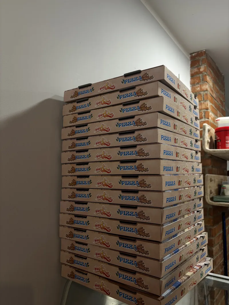 Una torre de cajas de pizza para llevar, de cartón kraft con «PIZZA» impreso en el canto, apiladas contra la pared blanca.