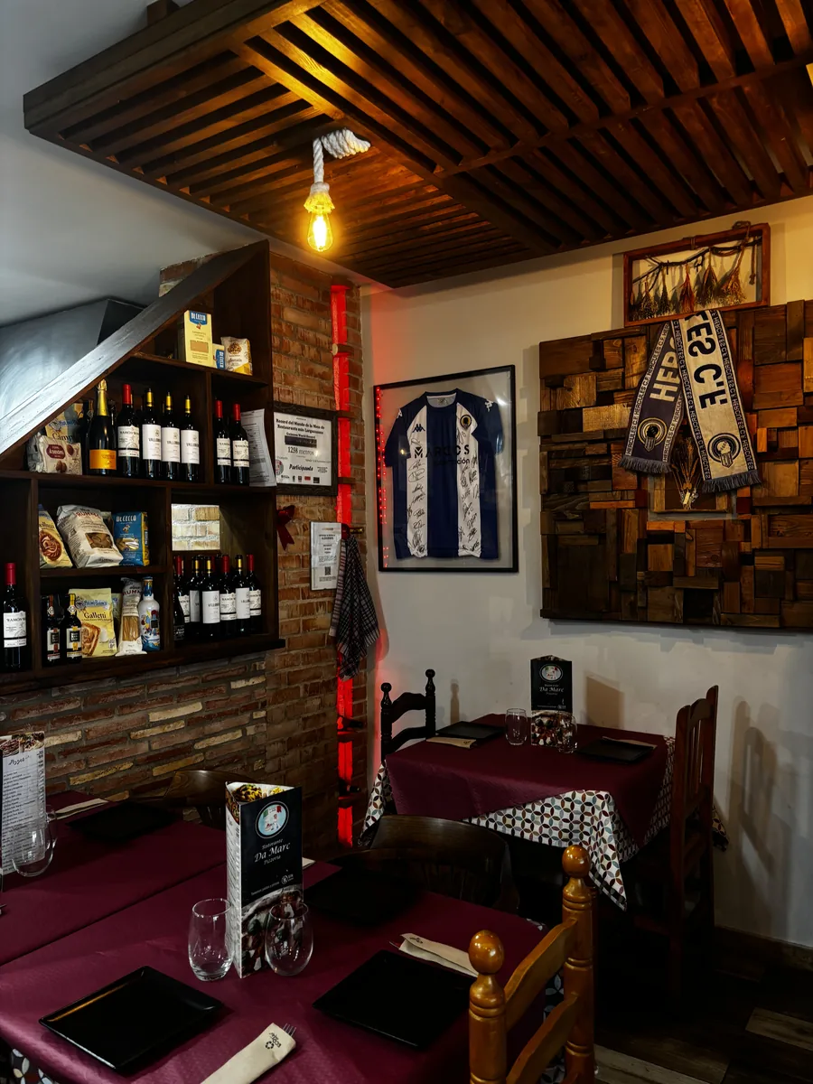 Nuestro comedor: mesas con mantel granate y platos negros cuadrados, sillas de madera torneada, una camiseta del Hércules enmarcada en la pared y la estantería con vinos y productos.