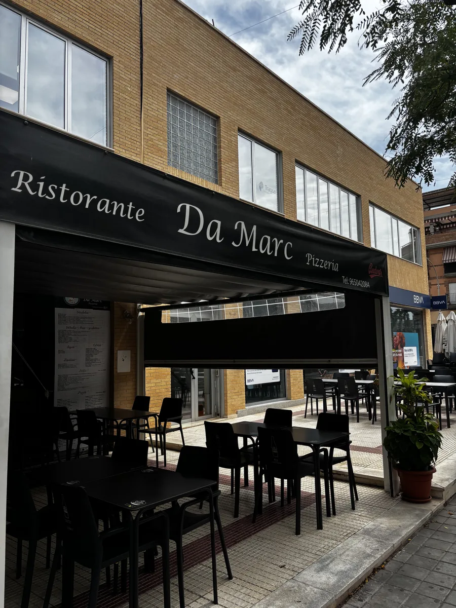 El toldo negro de nuestra fachada con el rótulo «Ristorante Da Marc Pizzería» y, debajo, las mesas y sillas negras de la terraza en la acera.